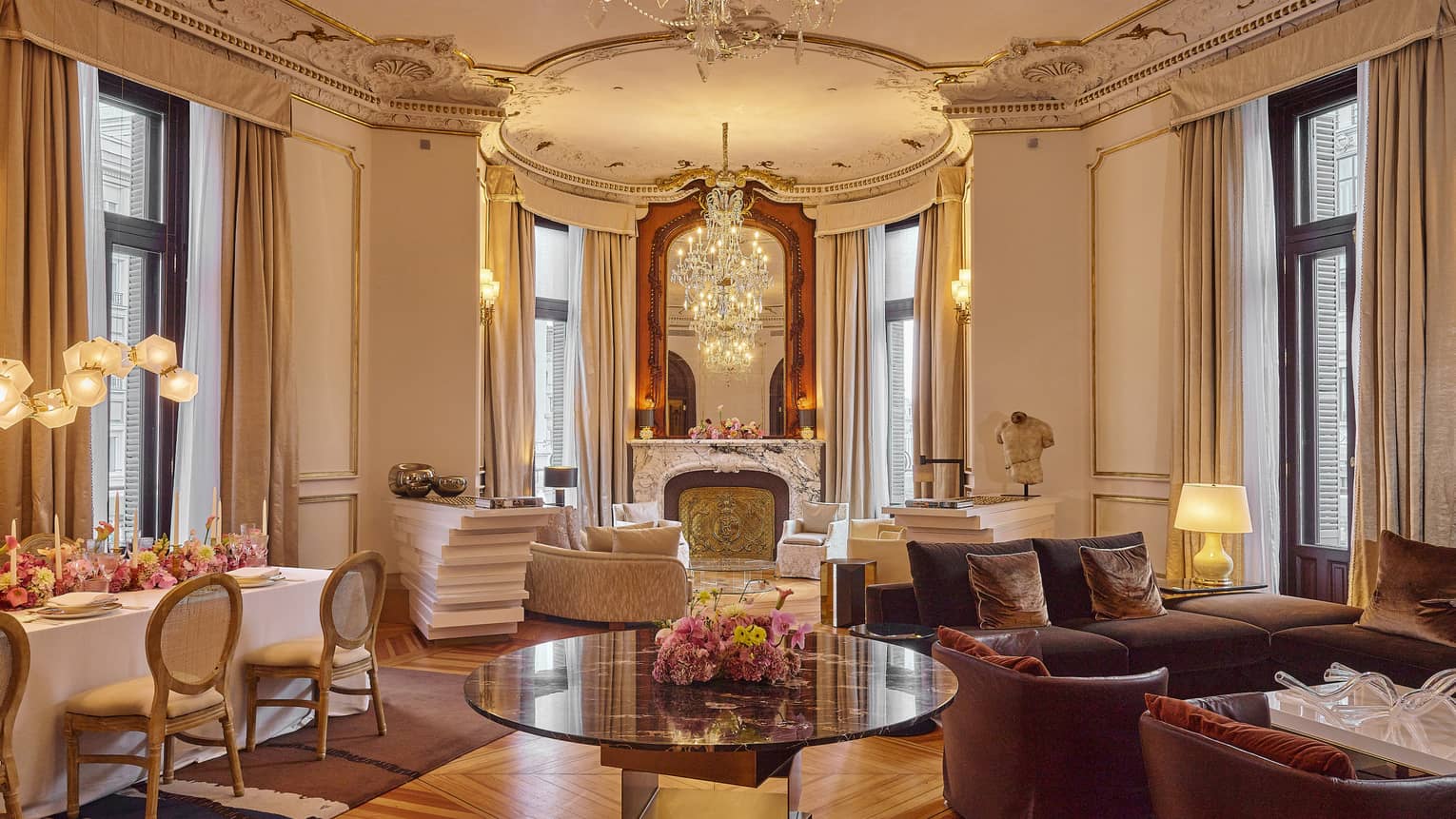 Large Regency-style living and dining area with tall ceilings, ornate finishes, two seating areas, a rectangular dining table and four sets of black-framed, floor-to-ceiling windows with beige curtains pulled to the side