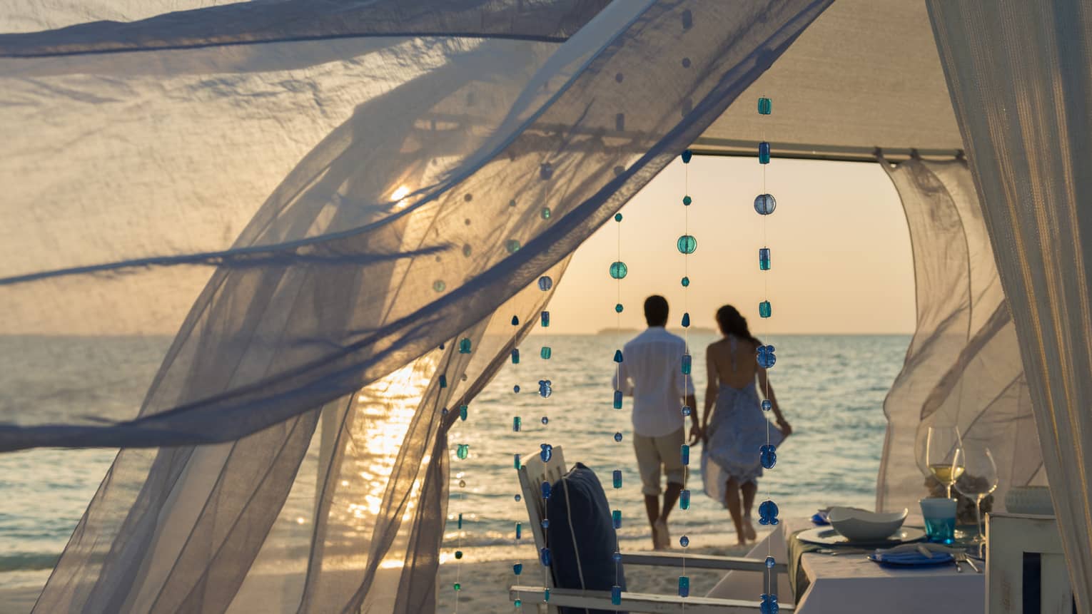 Sheer curtains blow in breeze, small blue beads hang from string, woman and man hold hands on beach in background