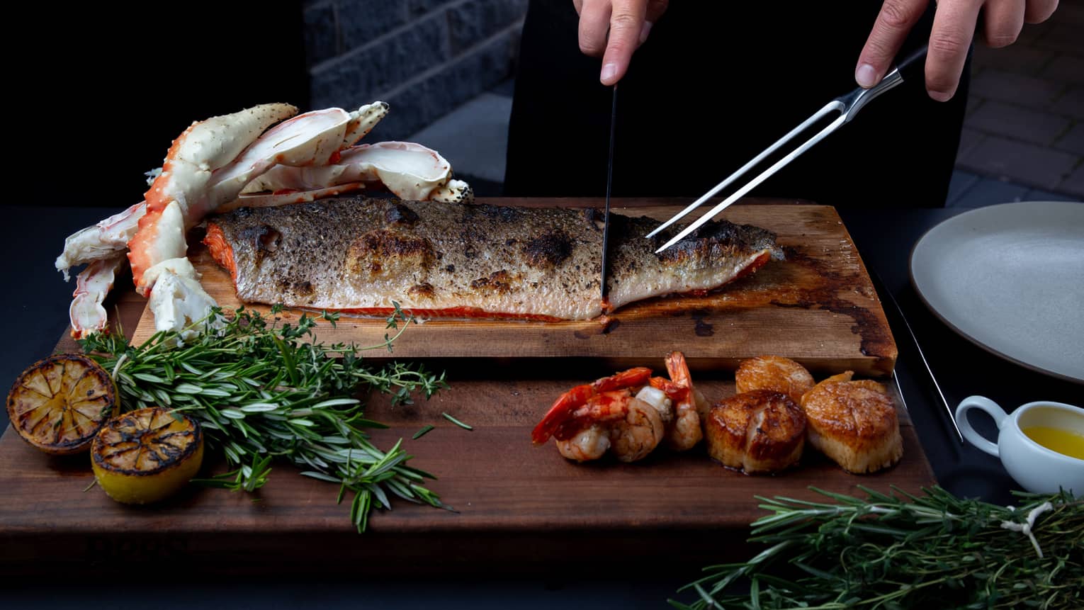 Closeup of chef preparing to carve a side of steelhead trout on a cedar plank, rosemary sprigs and charred lemon garnishes around
