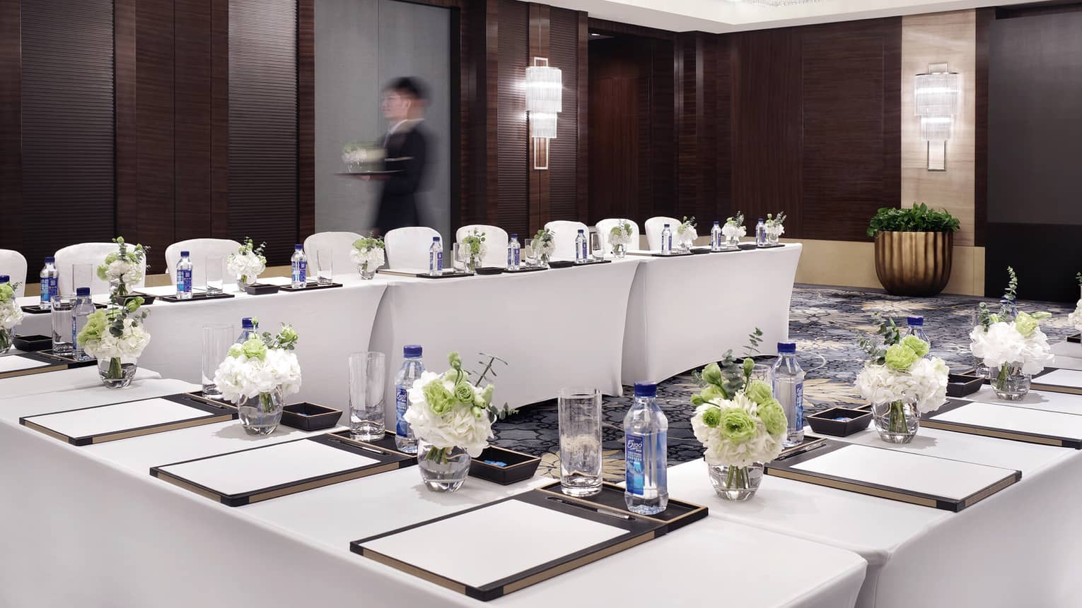 A person passes by carrying a tray of beverages in a modern conference room filled with banquet tables with writing pads, bottled water and floral arrangements at each seat