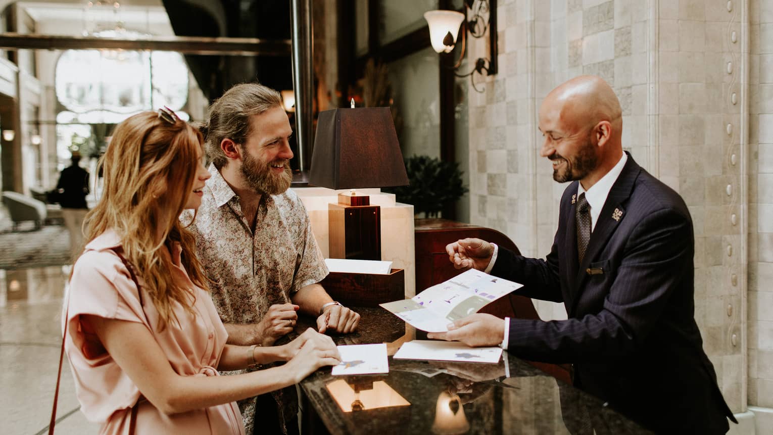 Couple at a hotel reception desk speaking with a Concierge, who is showing them a brochure.