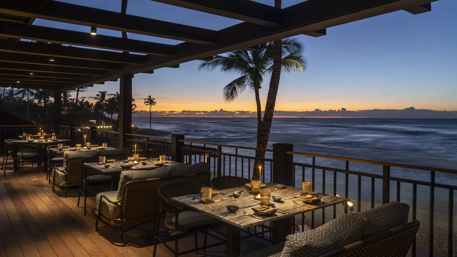 An intimately lit outdoor dining space with the ocean, palm trees and a dramatic sunset in the background