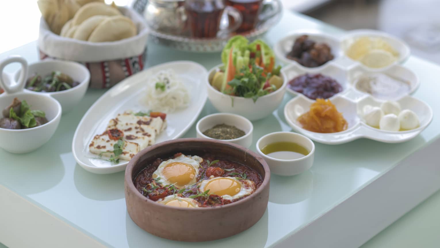 An assortment of traditional Arabic breakfast food on white platters, three eggs in a clay pot, pita in a basket and a glass tea tray