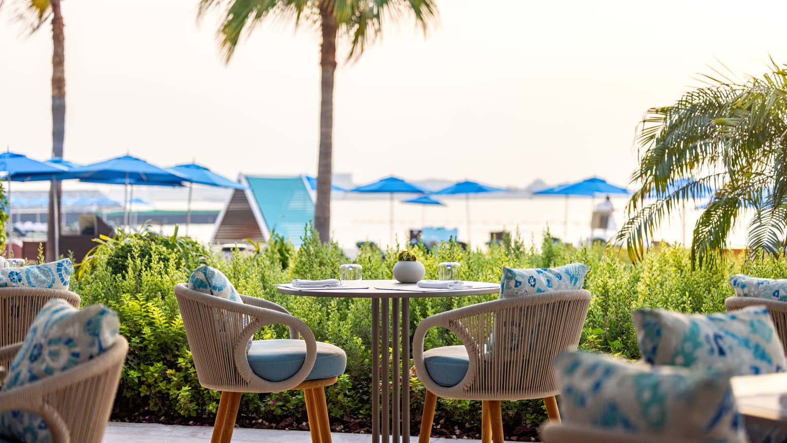 Small table for two set on an open-air terrace overlooking a lawn with palm trees and beach umbrellas in the distance