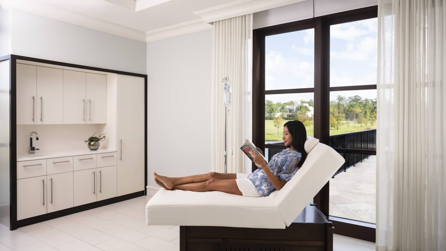 A woman reading a magazine on a folded mattress in a spa clinic with large window.