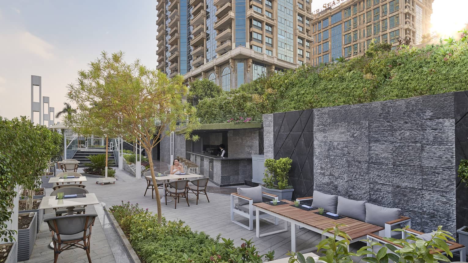 Woman seated at table on Riva restaurant concrete terrace amid greenery, hotel tower in backdrop