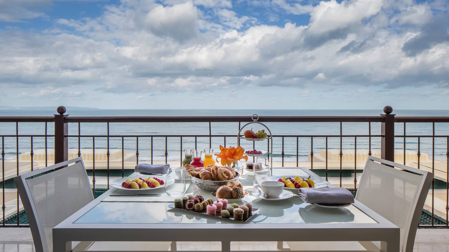 Balcony table set with fresh pastries, cakes, fruits, sea in background