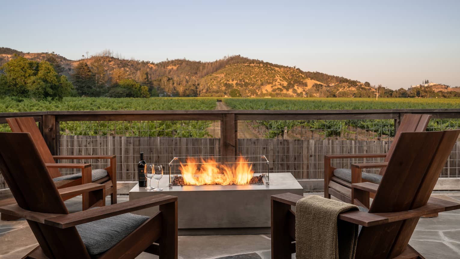 Four wooden chairs surround a bonfire on a stone terrace that overlooks vineyards