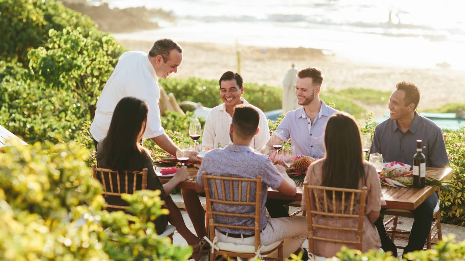 Six people smiling, sit around outdoor wooden table by ocean as wait staff serves them