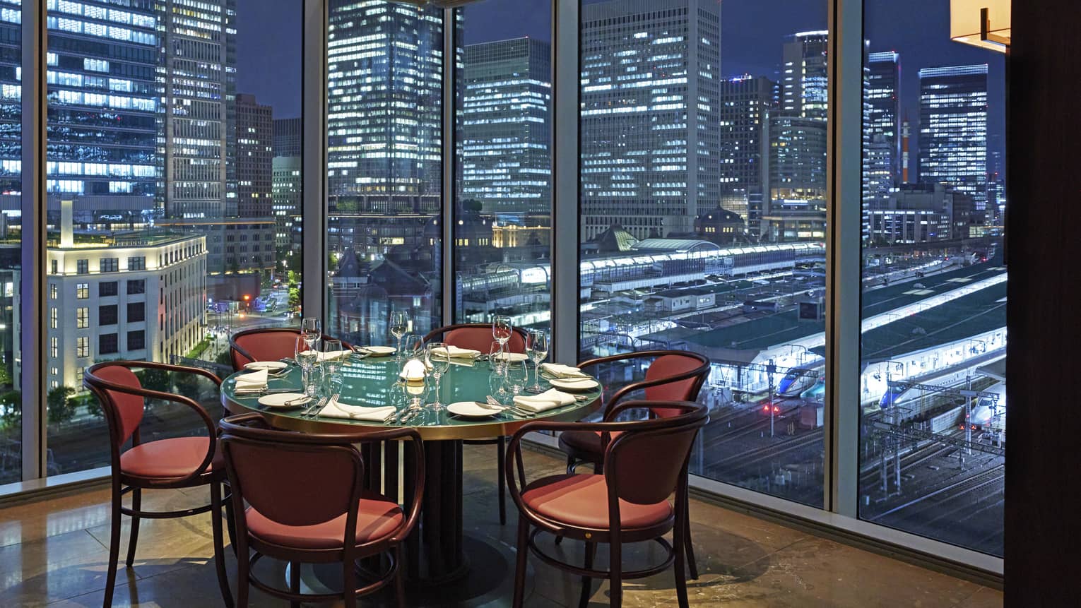 Luxury boutique hotel dining setup with a round table elegantly set for guests, offering a panoramic city view at night through tall glass windows