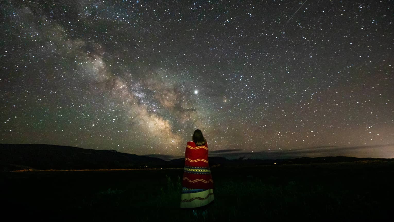 A lone woman stargazes, wrapped in a colourful striped blanket 