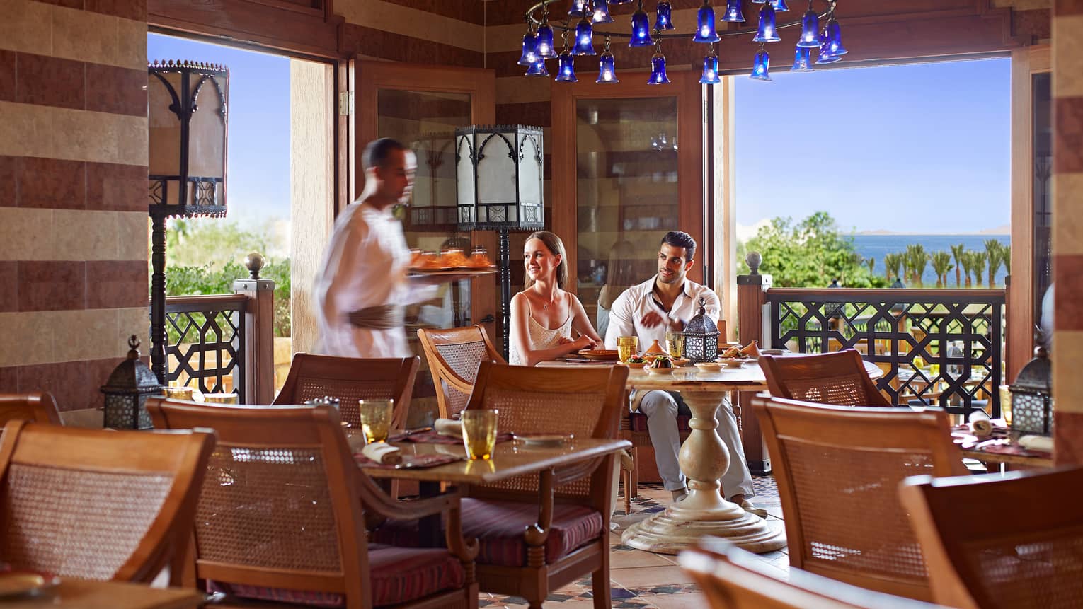 Couple sits at Arabesque corner table under blue chandelier, server walks by with tray
