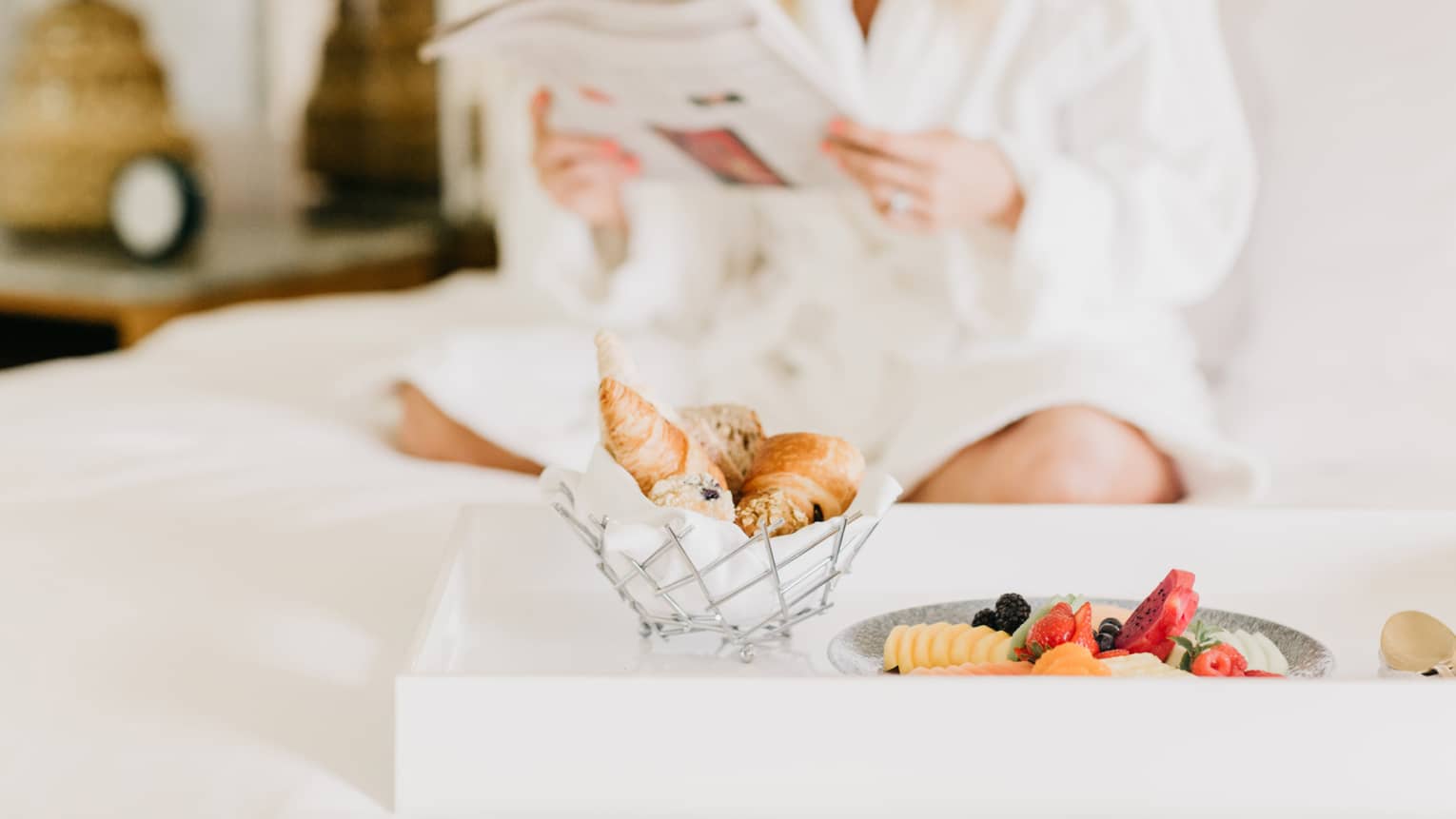 Pastries, fruit on in room dining tray, woman wearing white bath robe reads in background