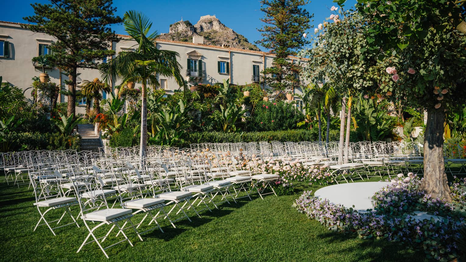 An outdoor event venue on a verdant lawn dotted with trees. Rows of white chairs are arranged in curved lines in front of a round dais framed by flowers.