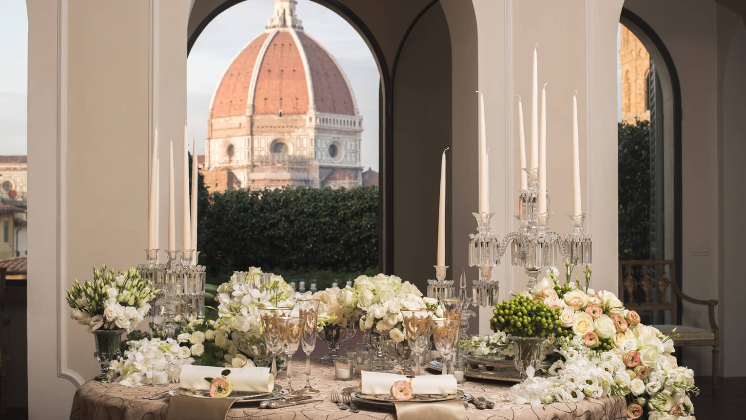 Elegant dining table set with candles, flowers, and glassware, with a large dome visible through arched windows in the background.