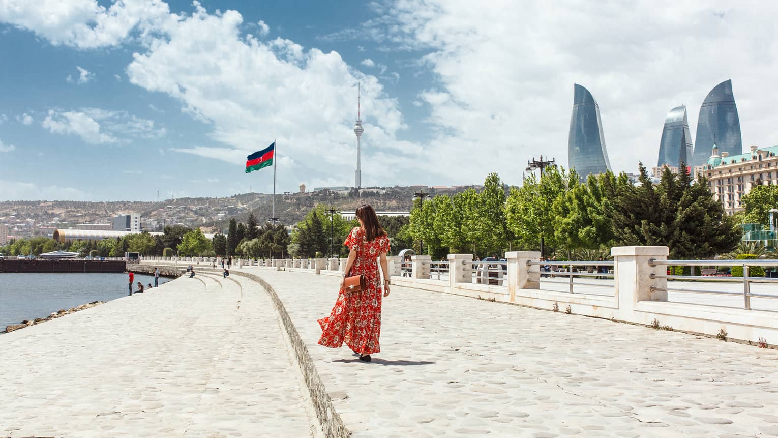 Woman wearing red dress walks along stone waterfront, skyscrapers in background
