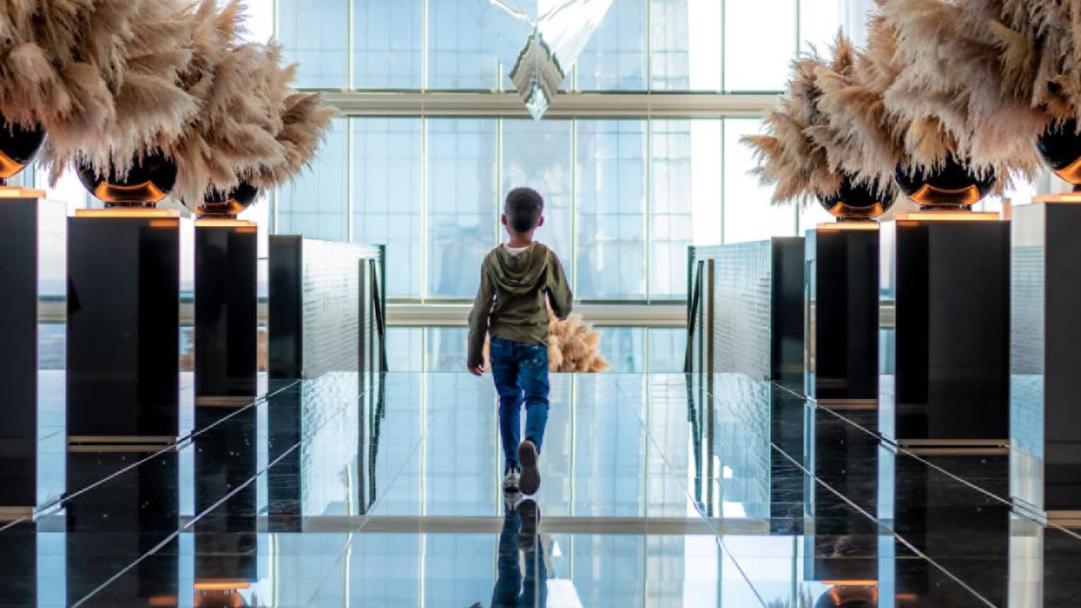 A small child in a hoodie walking in a room of glass walls and ceiling.