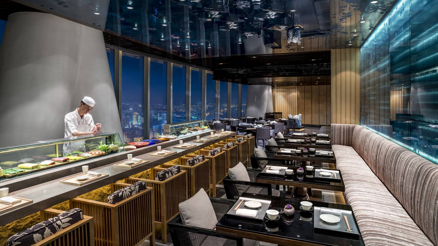 Kumoi Japanese restaurant chef stands behind long sushi bar in dining room overlooking city lights