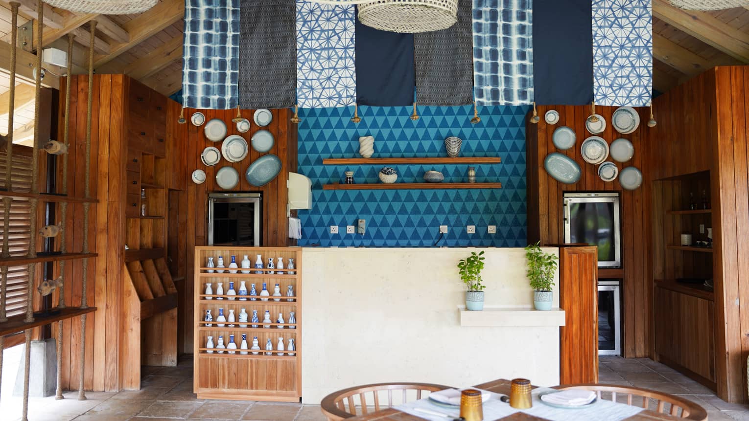 Casual restaurant with an island vibe, with blue and white fabric panels and rattan lights hanging from ceiling over teak and stucco bar
