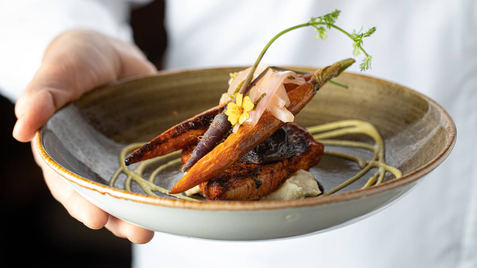 Chef holds dish with gourmet seared fish, carrots with pickled ginger, edible flowers