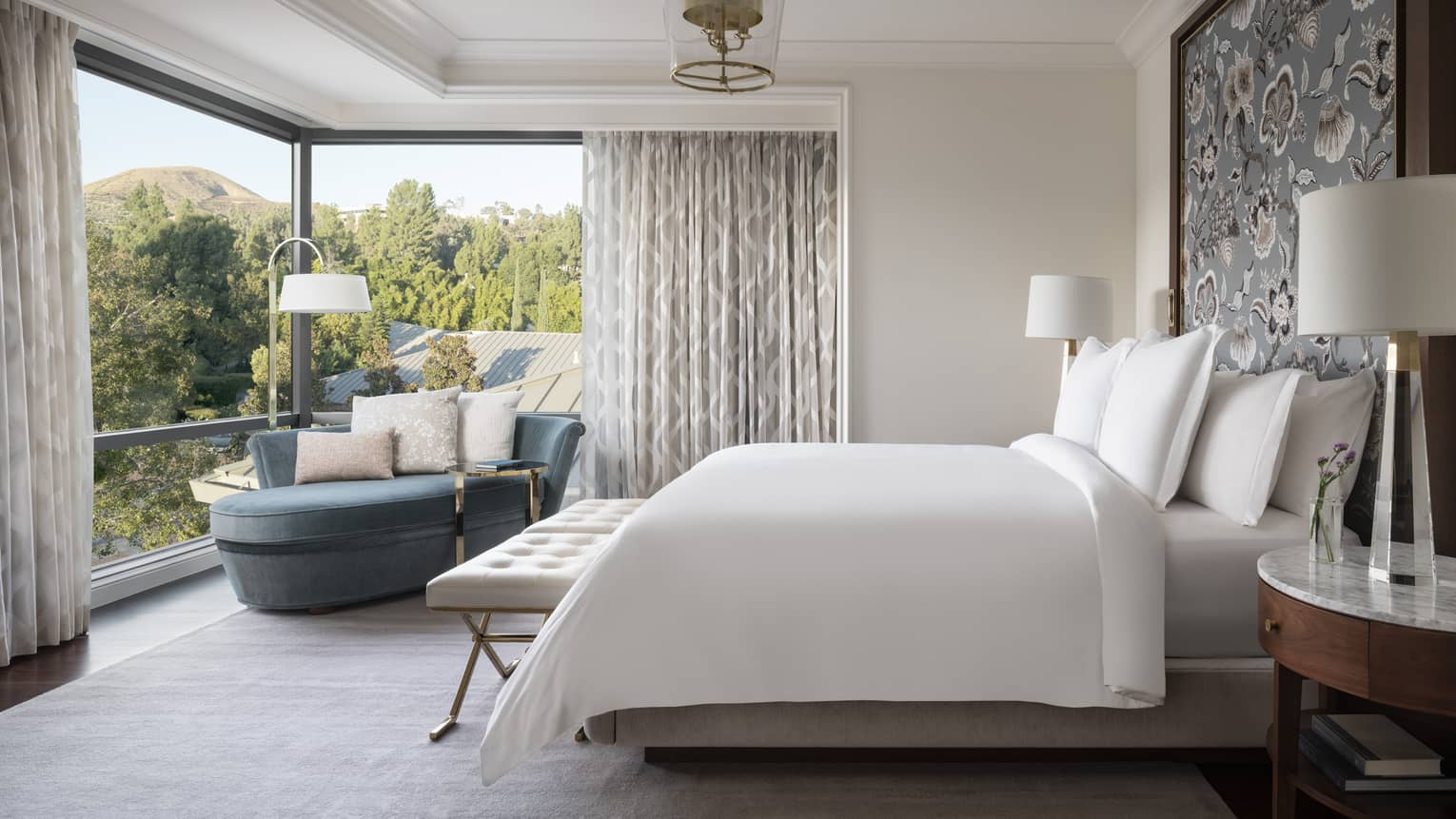 Guest room with white bed, blue chaise lounge, and a view of trees 