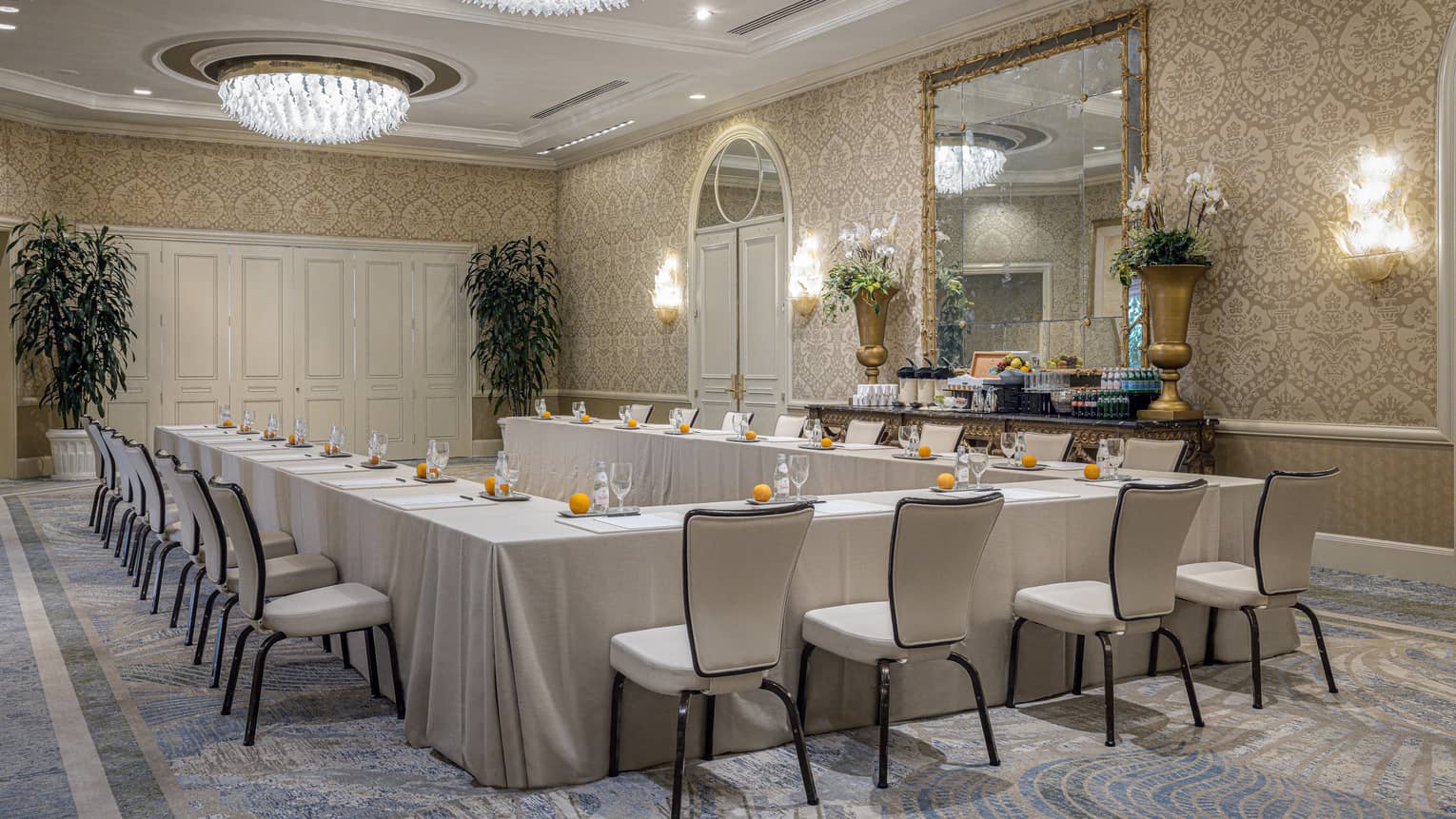 Meeting room set up hollow square with cream-coloured linens, dual chandeliers, lighted wall sconces and large mirror on patterned wall