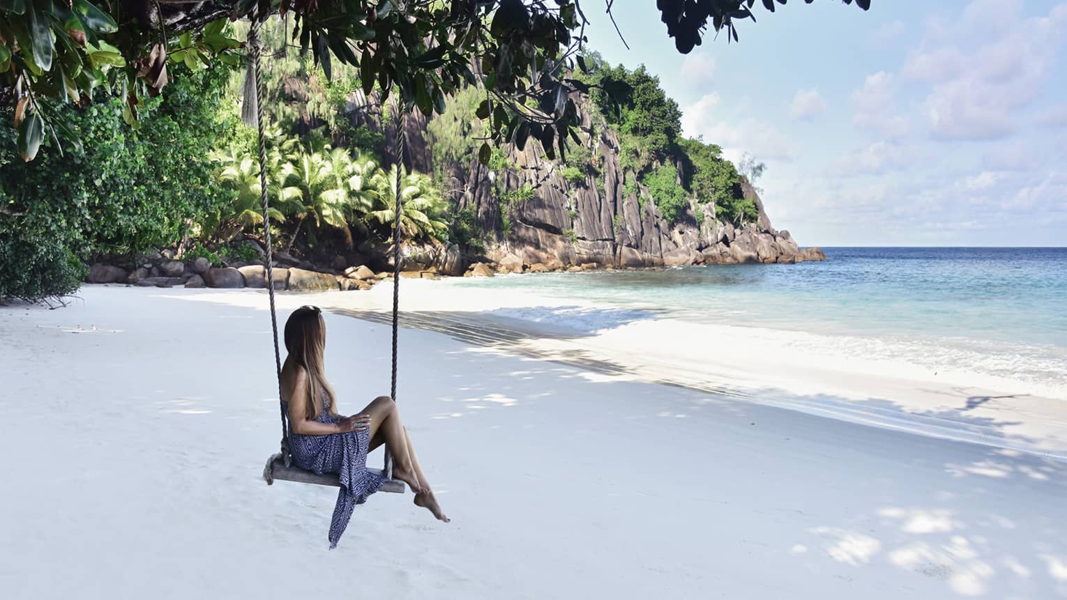 A woman sits on a tree swing on the beach with a view of a cliffside and the ocean