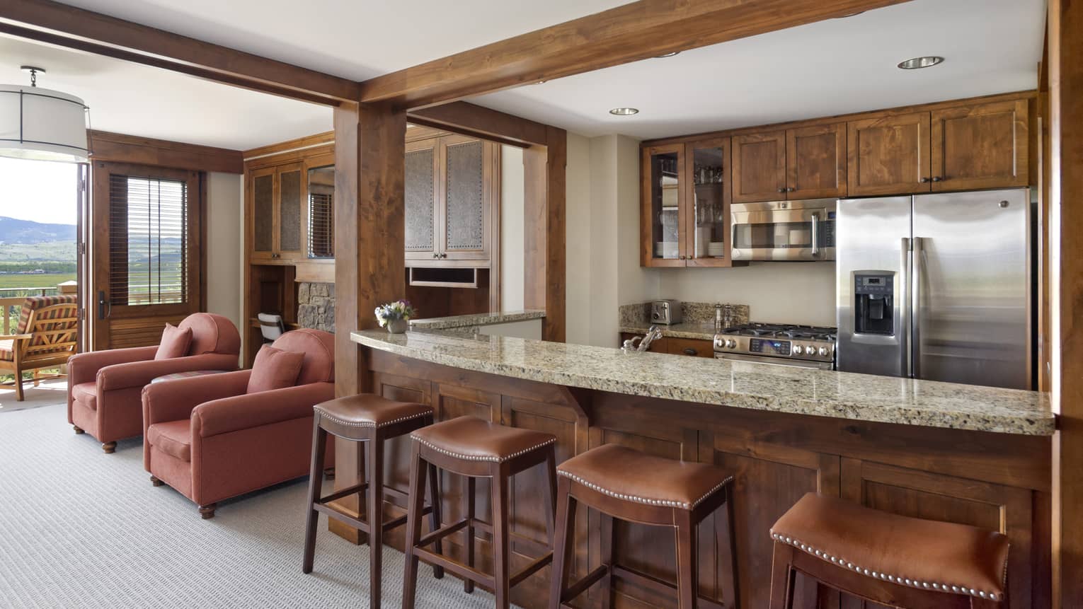 Kitchen counter with four stools, wooden cabinetry, stainless steel appliances, and two red armchairs near a balcony with a mountain view