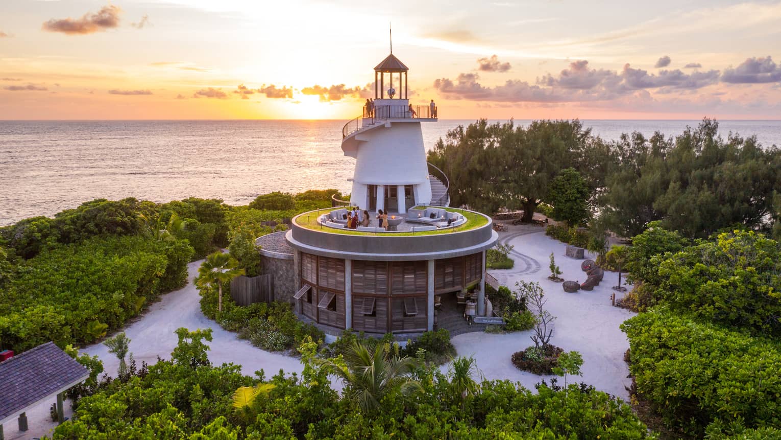 Lighthouse nestled among greenery with people on different levels, overlooking the ocean at sunset