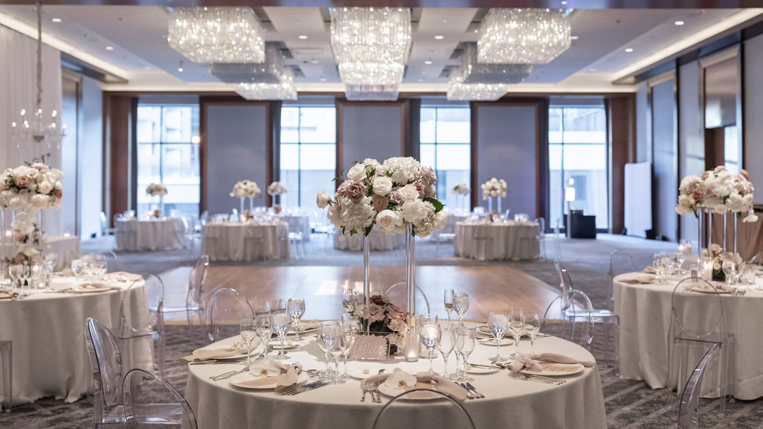 A well lit ballroom with ground tables and flower centerpieces and a section of hardwood floor in the middle.