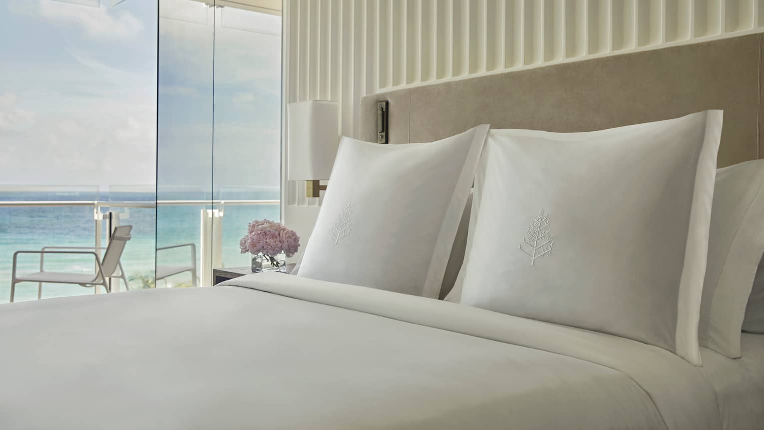 Luxury hotel bed with white embroidered pillows, a bedside table with flowers and floor-to-ceiling windows offering ocean views