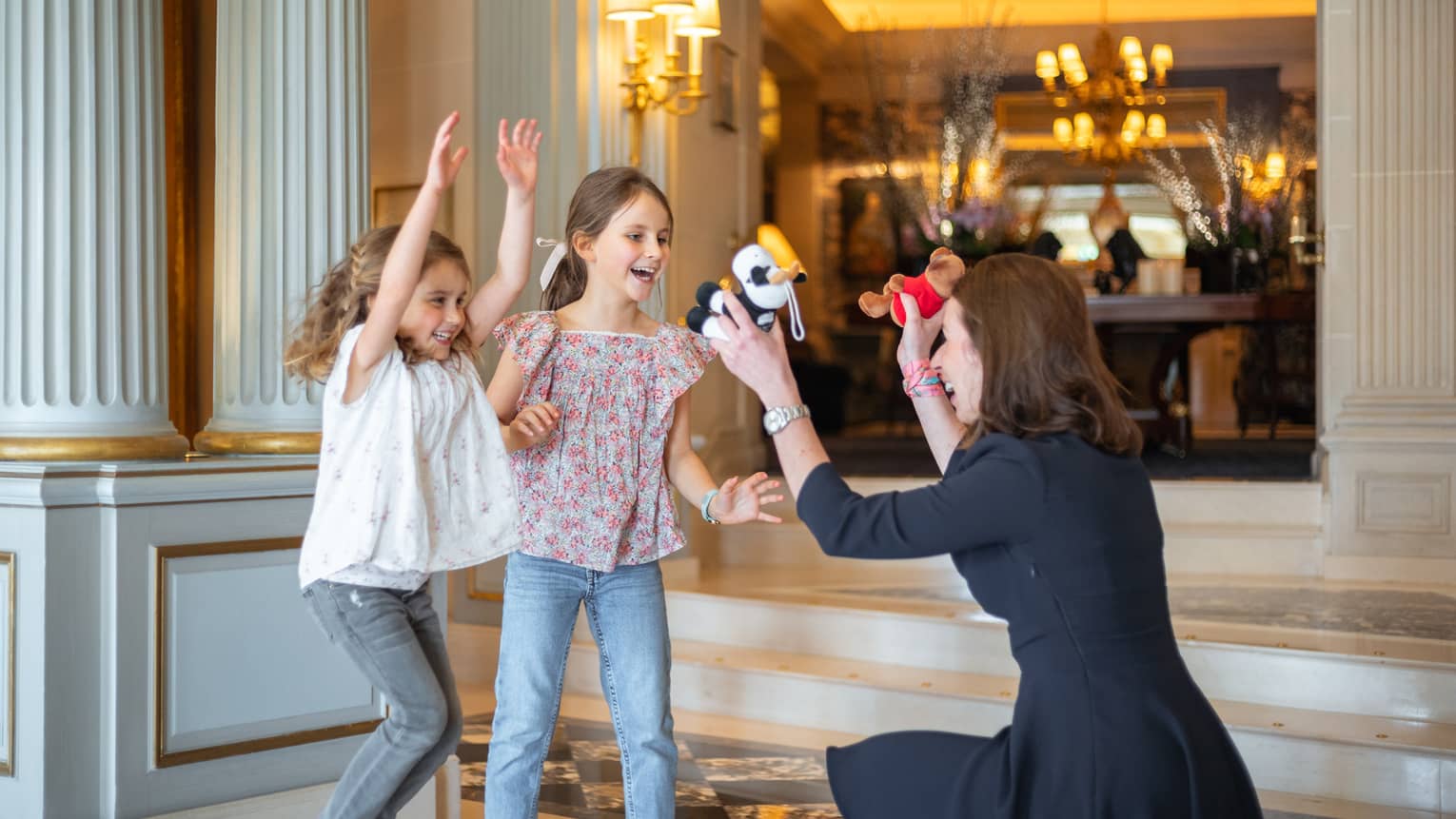 Concierge crouched, holding two stuffed animals toward excited, smiling girls