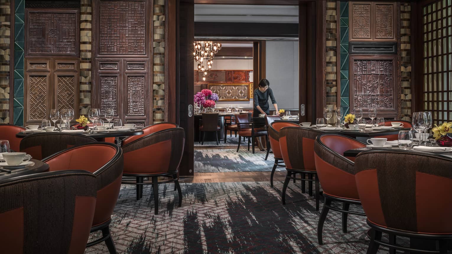 Woman sets table at Jiang-Nan Chun, Michelin-starred Cantonese restaurant, with red leather bucket chairs around dining tables, carved wood walls.