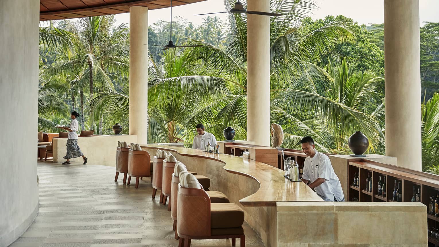 Smiling bartenders behind long wood-and-stone bar under white pillars of resort balcony, palm trees in background