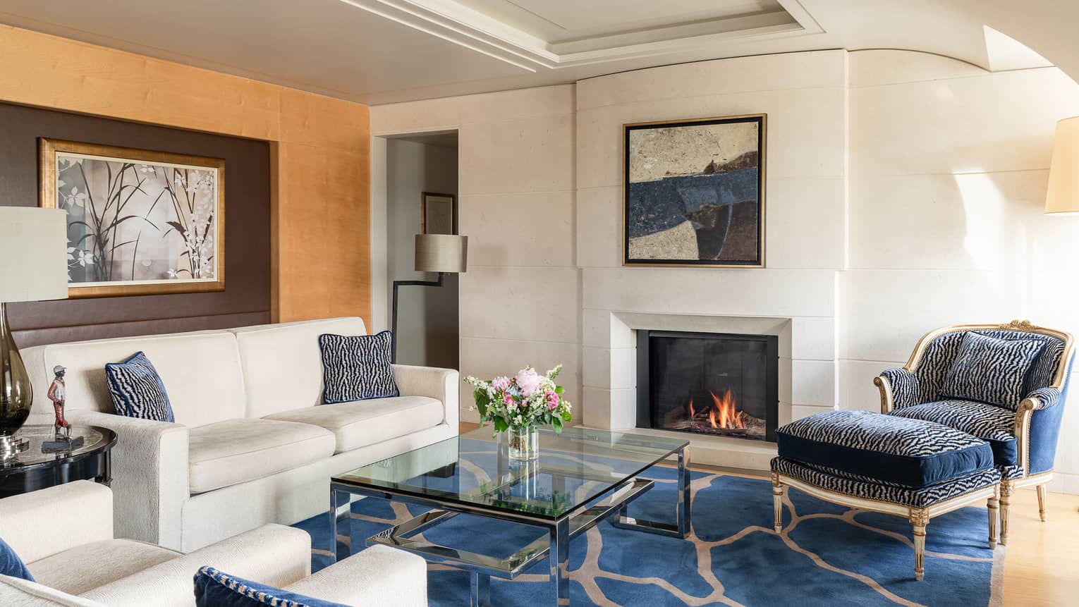 Luxury hotel suite living room with a cosy fireplace, cream sofa, blue and cream armchairs, glass coffee table, floral arrangement and wall art