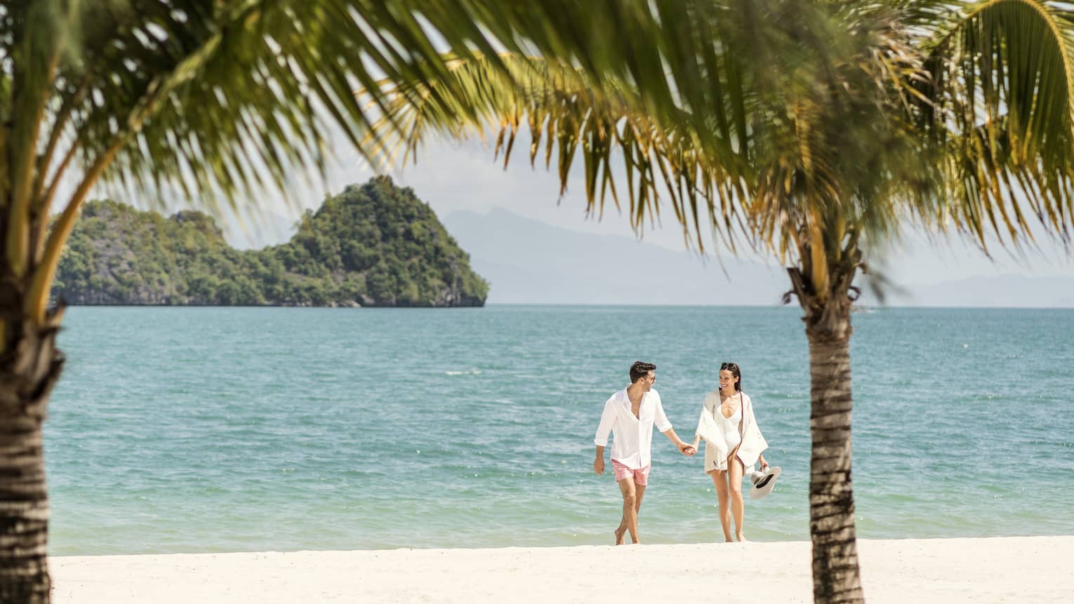 A man and woman dressed in white, holding hands while walking on the beach