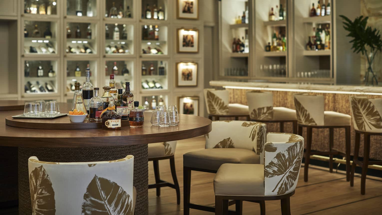 A round wood table set for a rum tasting in an elegant bar, backlit shelves around the room filled with bottles and glasses.