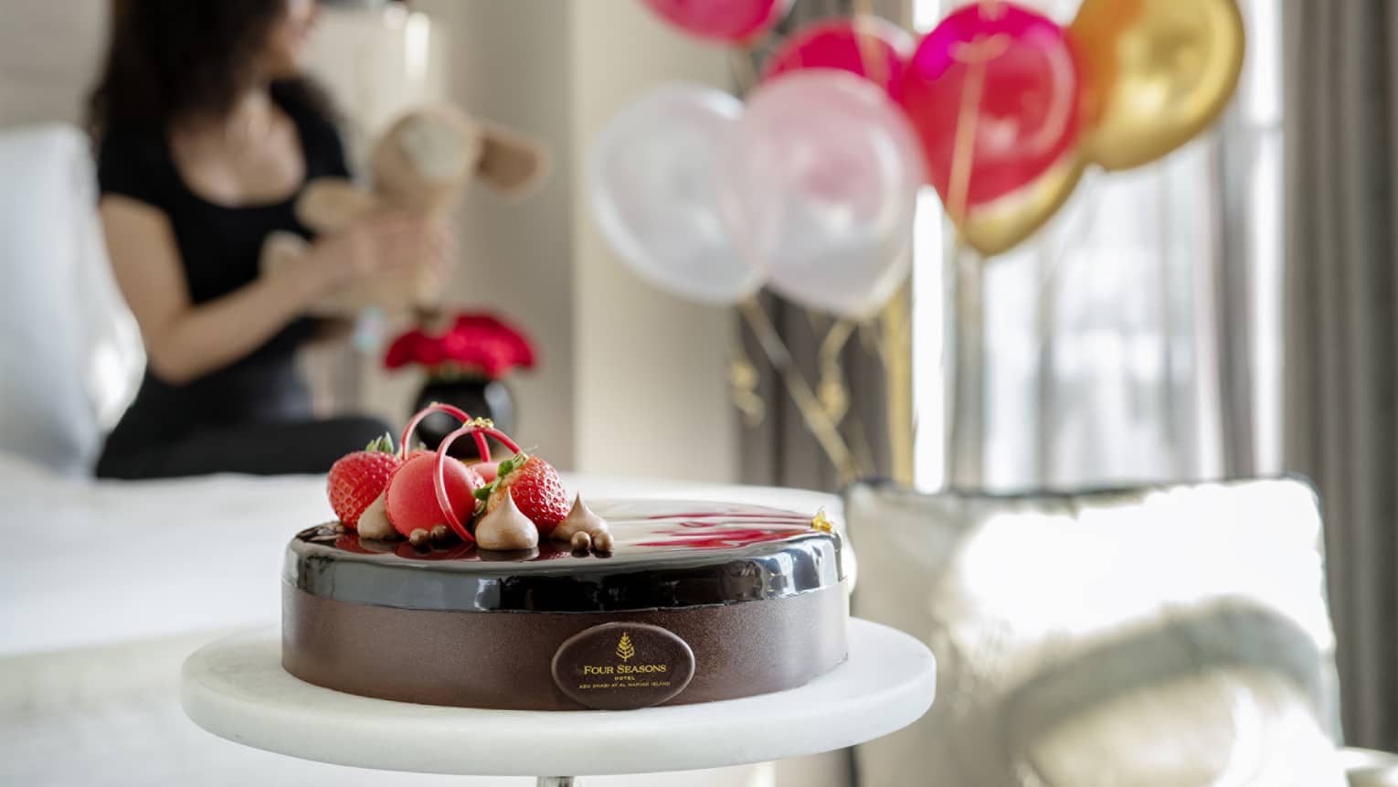 Chocolate cake topped with candies and red berries sits in the centre of a hotel guest room with a person wearing a black dress and holding a stuffed animal sits on the bed behind it next to a bouquet of pink, gold and white balloons