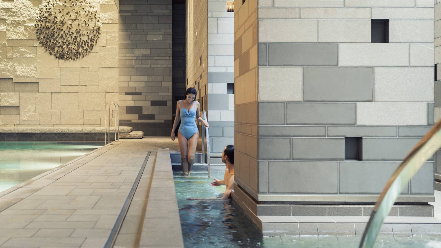 A guest enters a pool via the stairs to meet another guest who is half-hidden by a large grey-tiled column in the foreground.