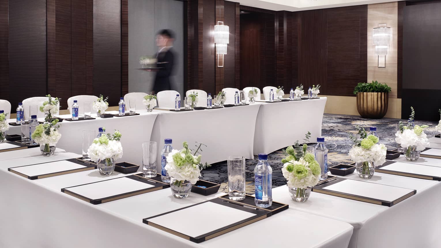 A person passes by carrying a tray of beverages in a modern conference room filled with banquet tables with writing pads, bottled water and floral arrangements at each seat