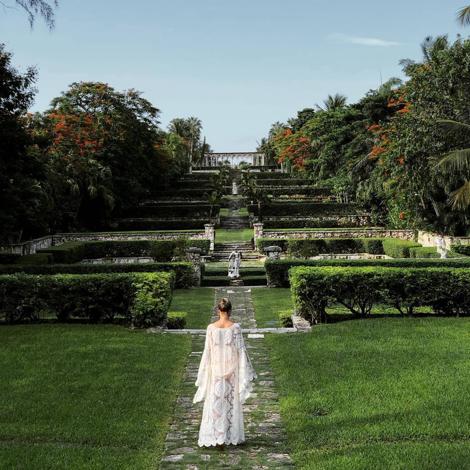 Person in a white flowing dress walking through terraced gardens with stone paths and lush greenery