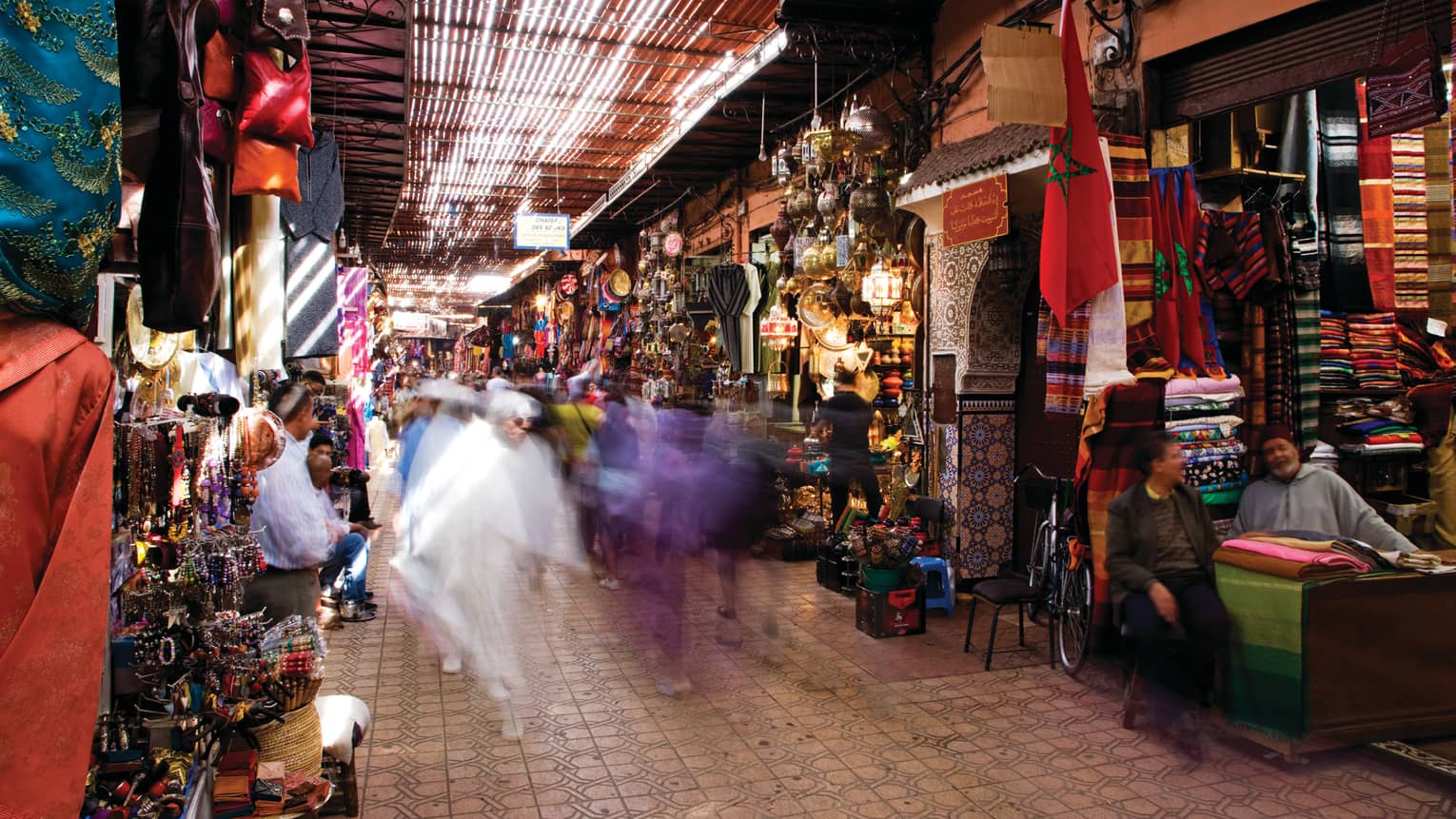 People walk through Moroccan market with stalls filled with colourful textiles, treasures
