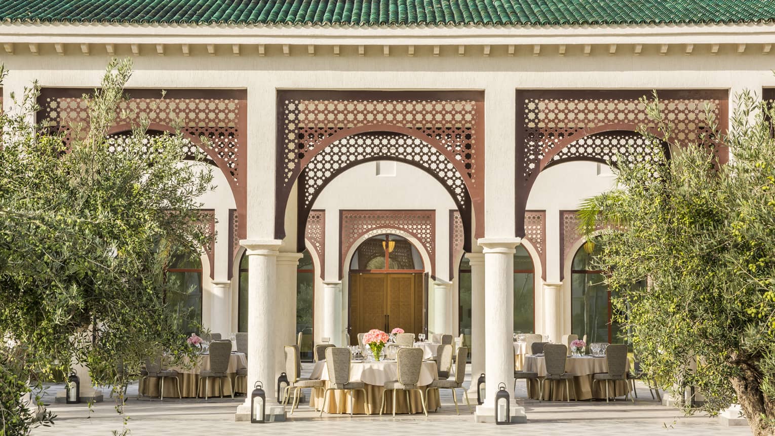 Round tables and chairs with vases of flowers set on covered patio with trees, lanterns 