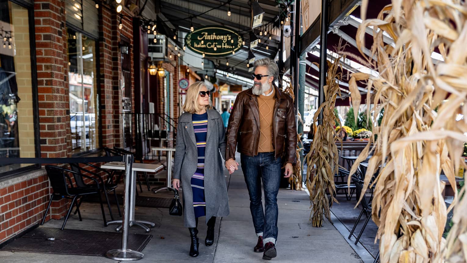 A man and woman walking by a brick building with sunglasses and warm clothes on.