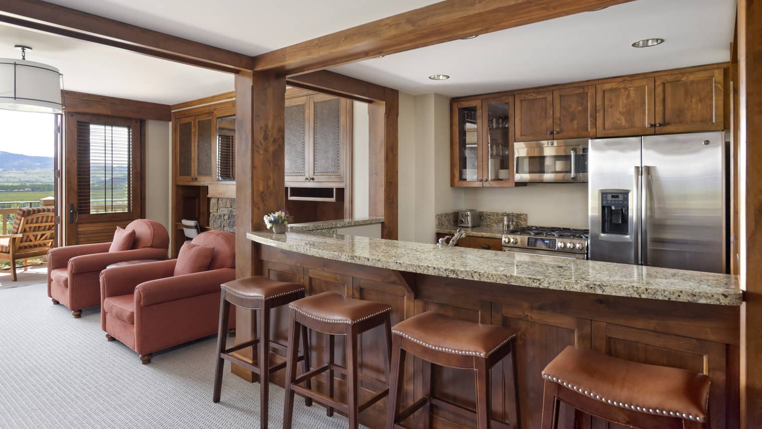 Kitchen counter with four stools, wooden cabinetry, stainless steel appliances, and two red armchairs near a balcony with a mountain view