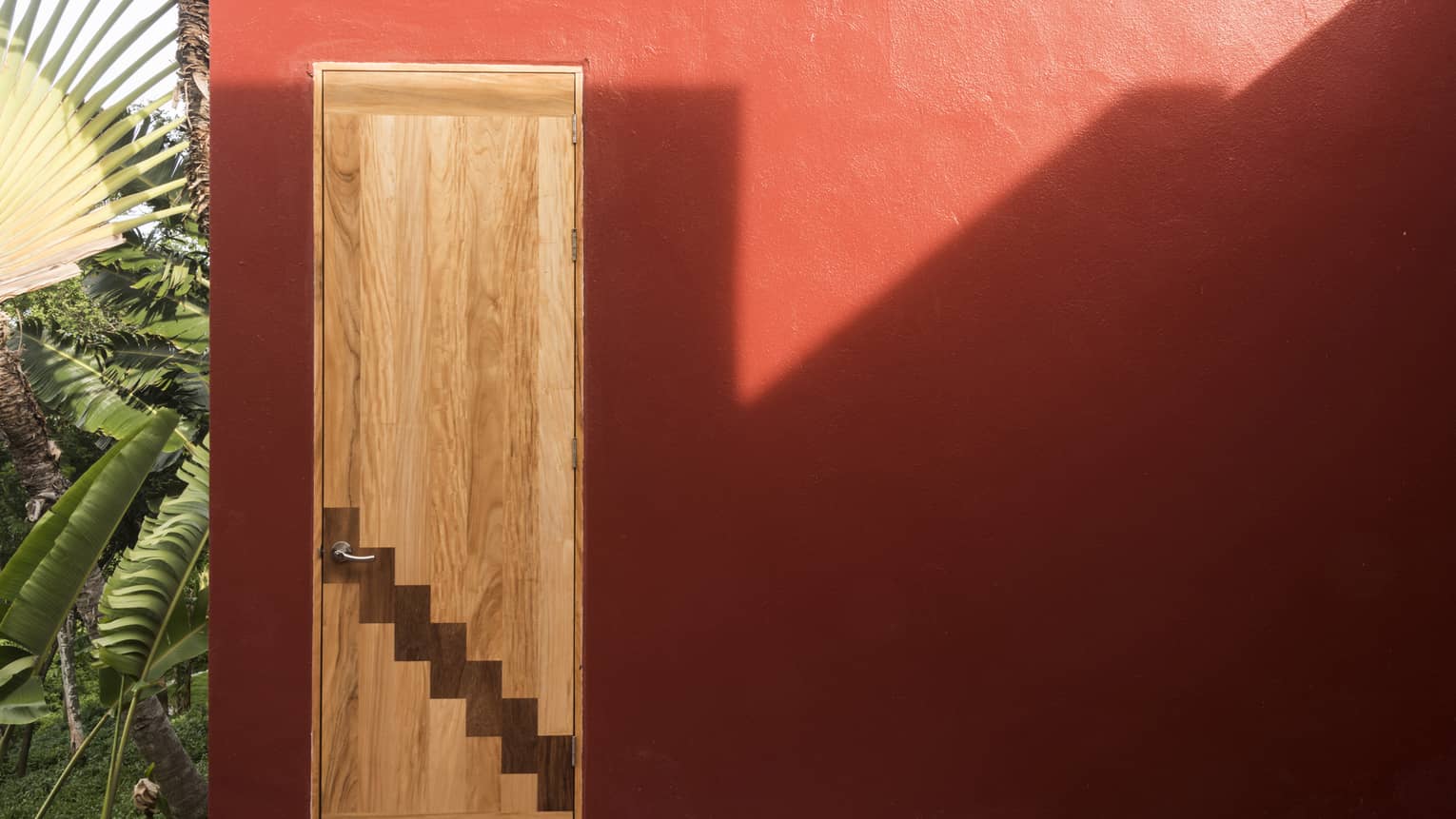 Red wall of a villa with a contemporary wooden door and palm trees
