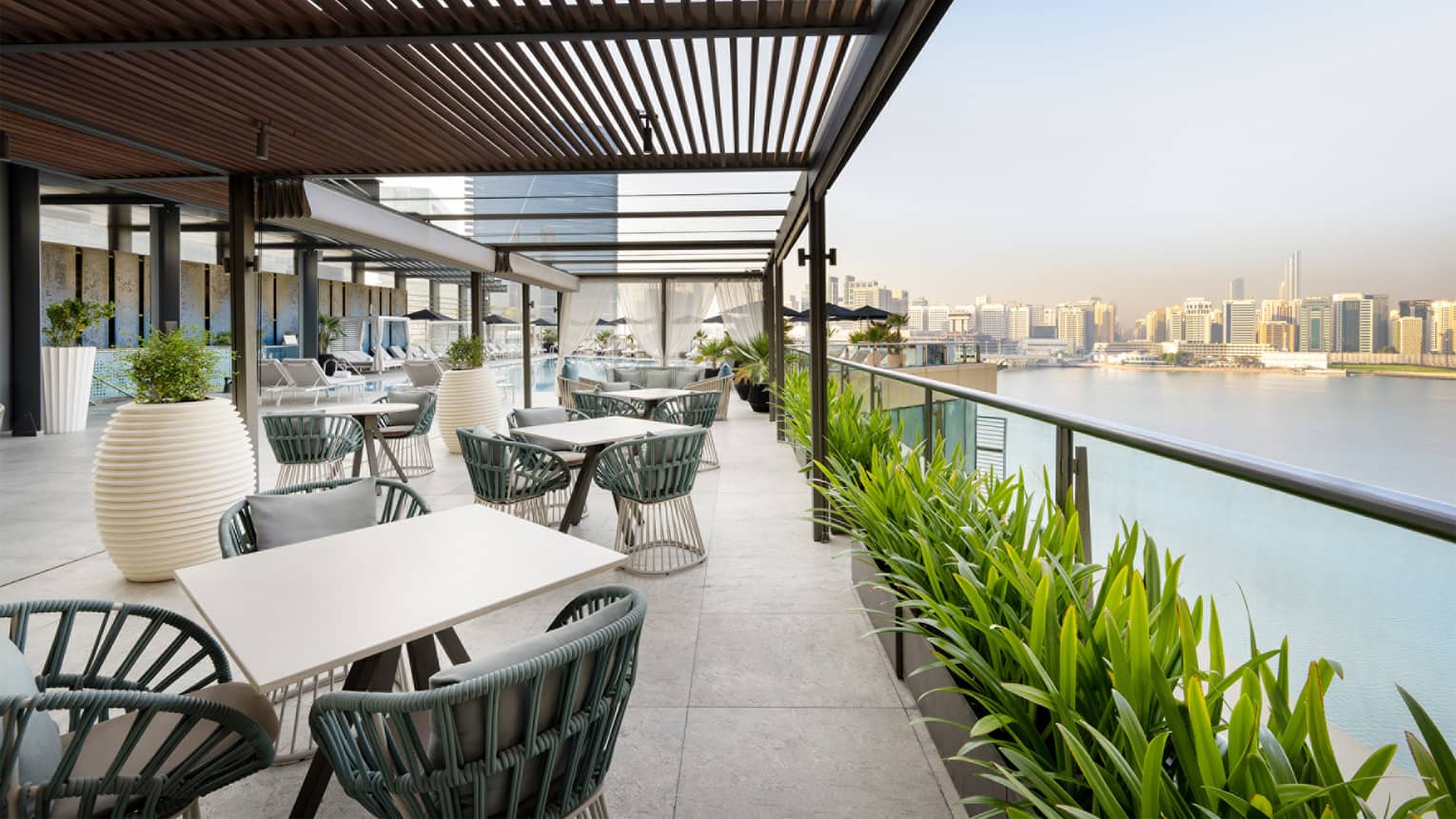 Restaurant terrace with slated wood overhang, various white tables, dark-teal chairs and a row of green plants overlooks the water with a city skyline in the background
