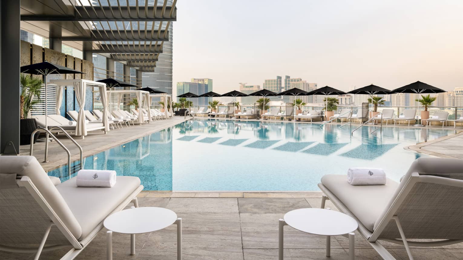 Outdoor pool surrounded by white chairs, a row of cabanas, a row of black umbrellas and a city skyline in the distance