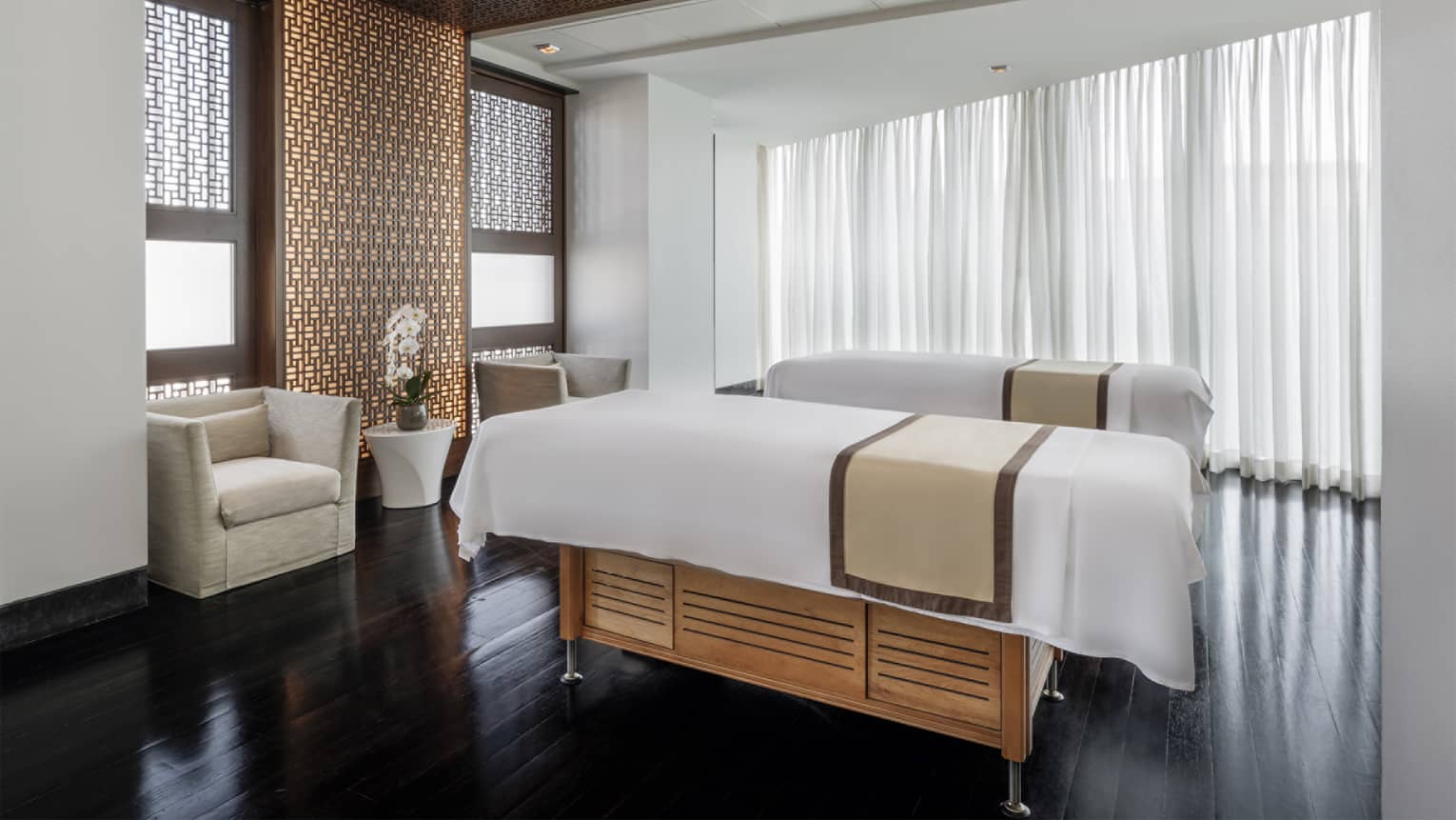 Couples' spa treatment room with two white-linen-covered massage tables, two arm chairs, a small table with flowers and a wall of curtain-covered windows along with two smaller windows facing the tables