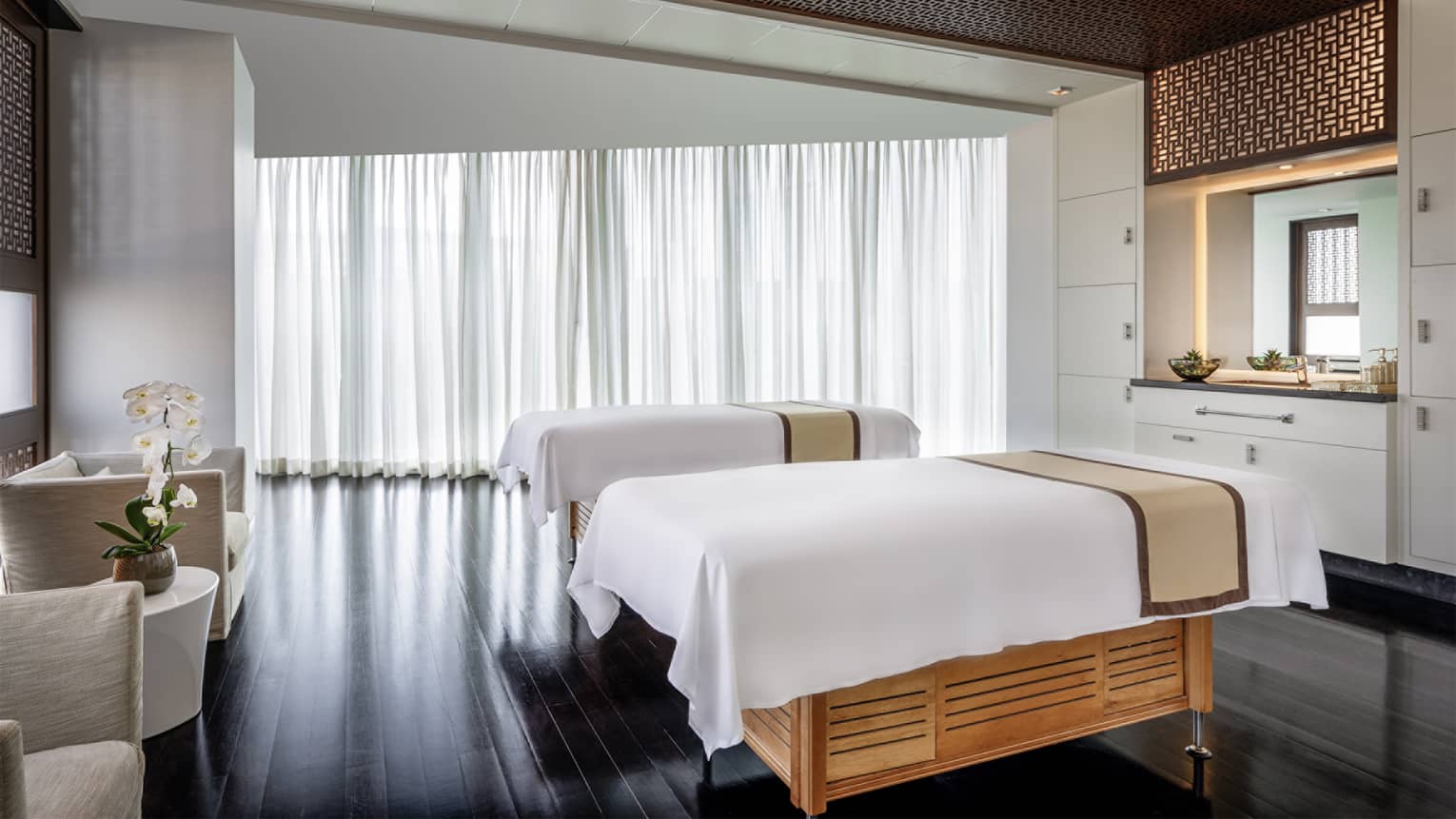 Large spa treatment room with two white-cloth-covered massage tables, two linen arm chairs, a vanity area, wall of windows draped in white curtains with the sun shining through and dark-wood flooring